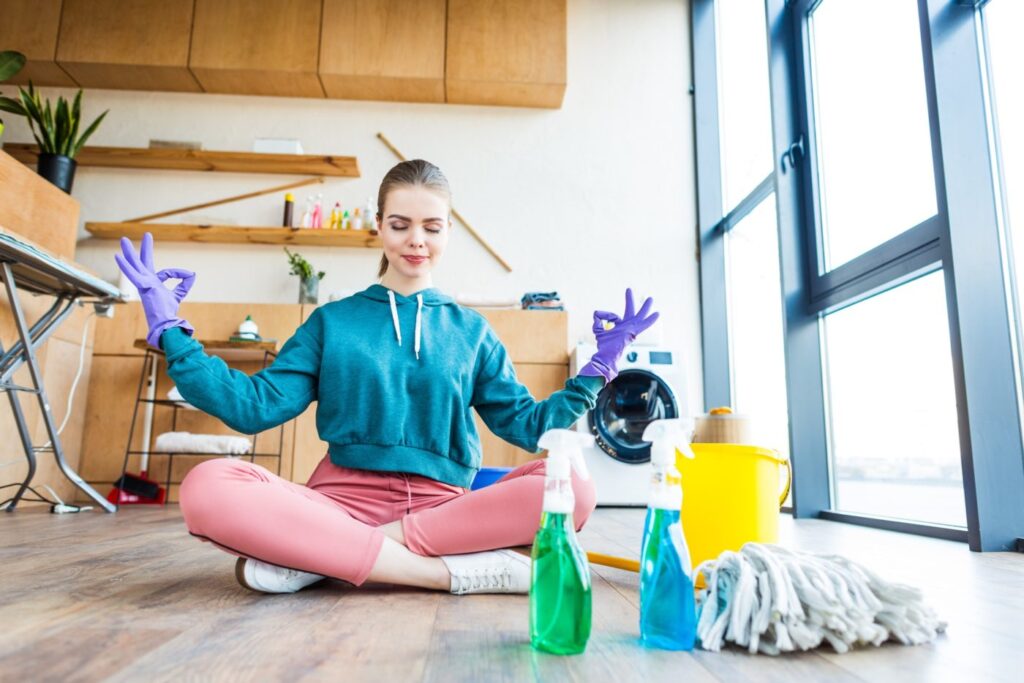 smiling young woman sitting on floor and meditating while cleaning house e1669624929148