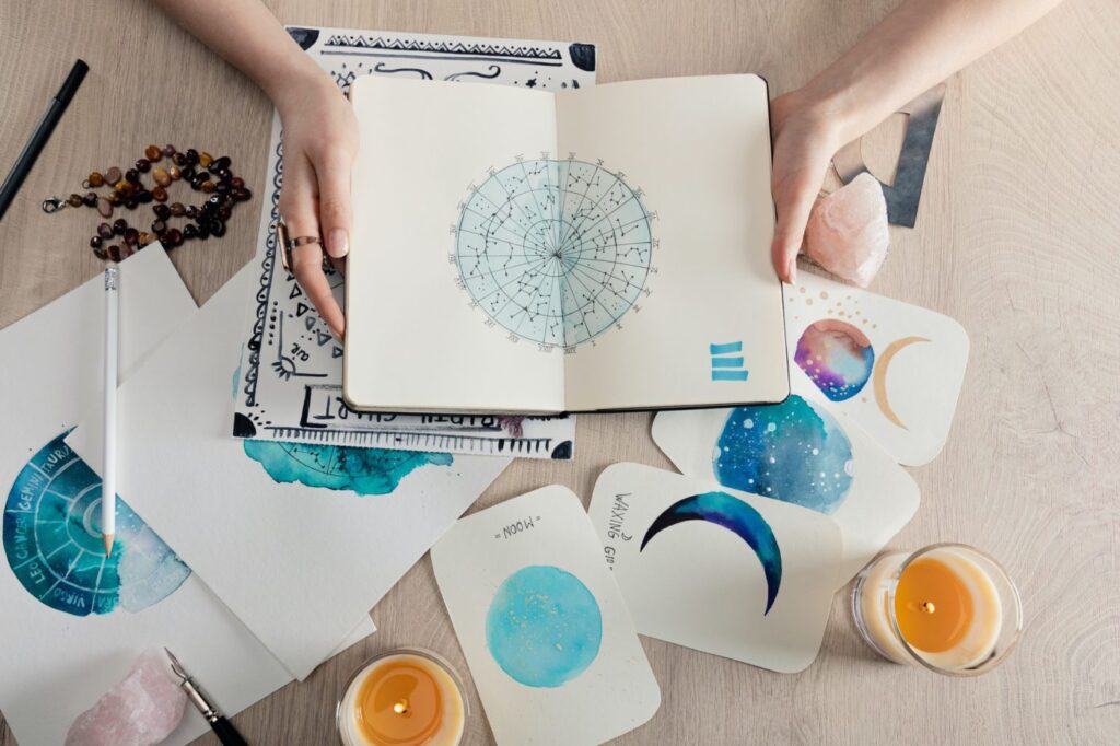 top view of astrologer holding notebook with watercolor drawings and zodiac signs on cards on table e1669624941145